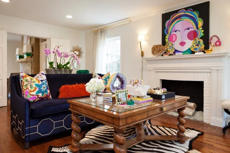 Living room with dark blue sofa, colorful floral cushions, orange cushion, wooden table, striped rug and bright portrait artwork over a white fireplace.
