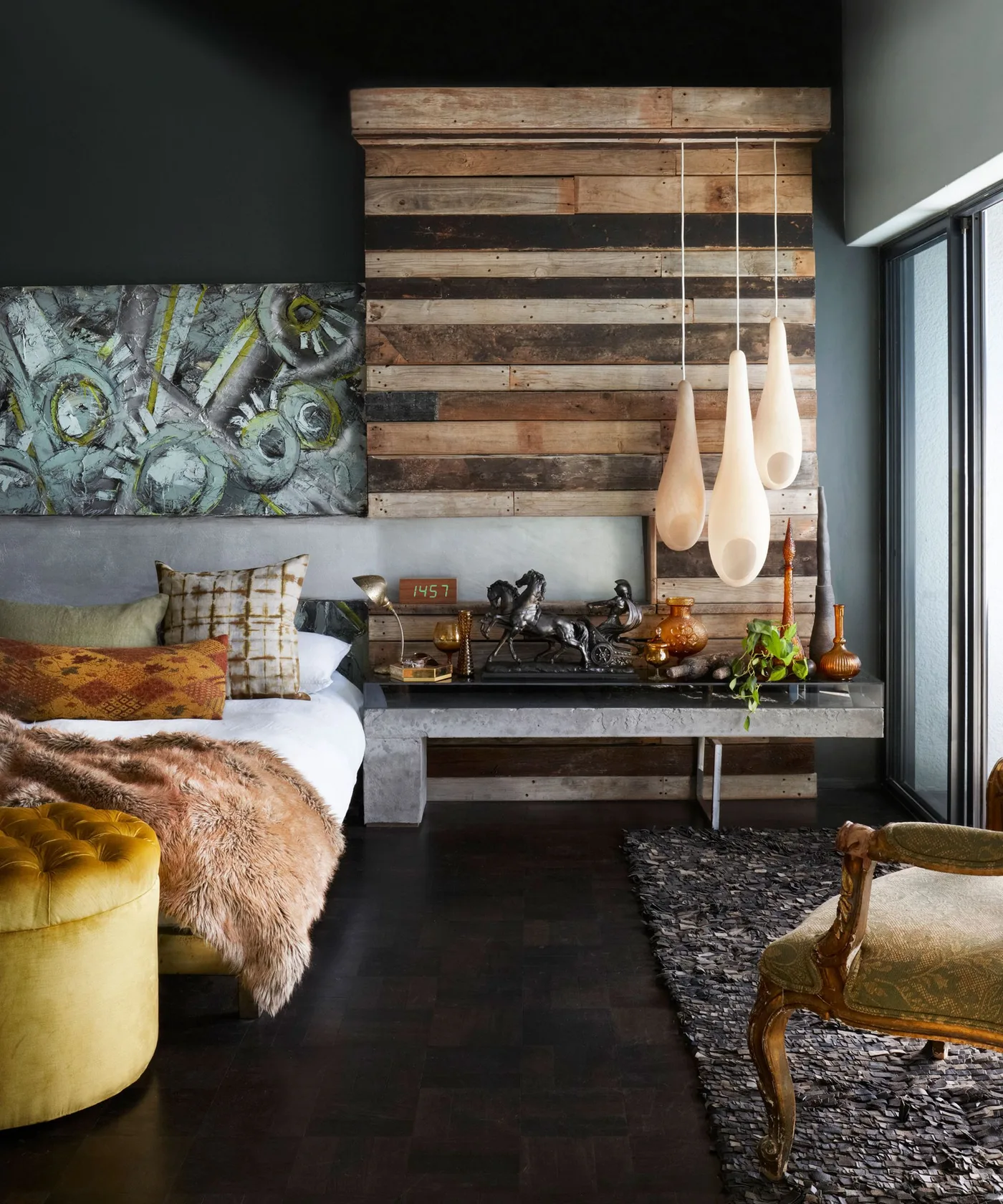 Dark bedroom with horizontal wooden wall boards, pale elongated pendant lights, textured bed throws, yellow pouf and curved wooden-framed chair.