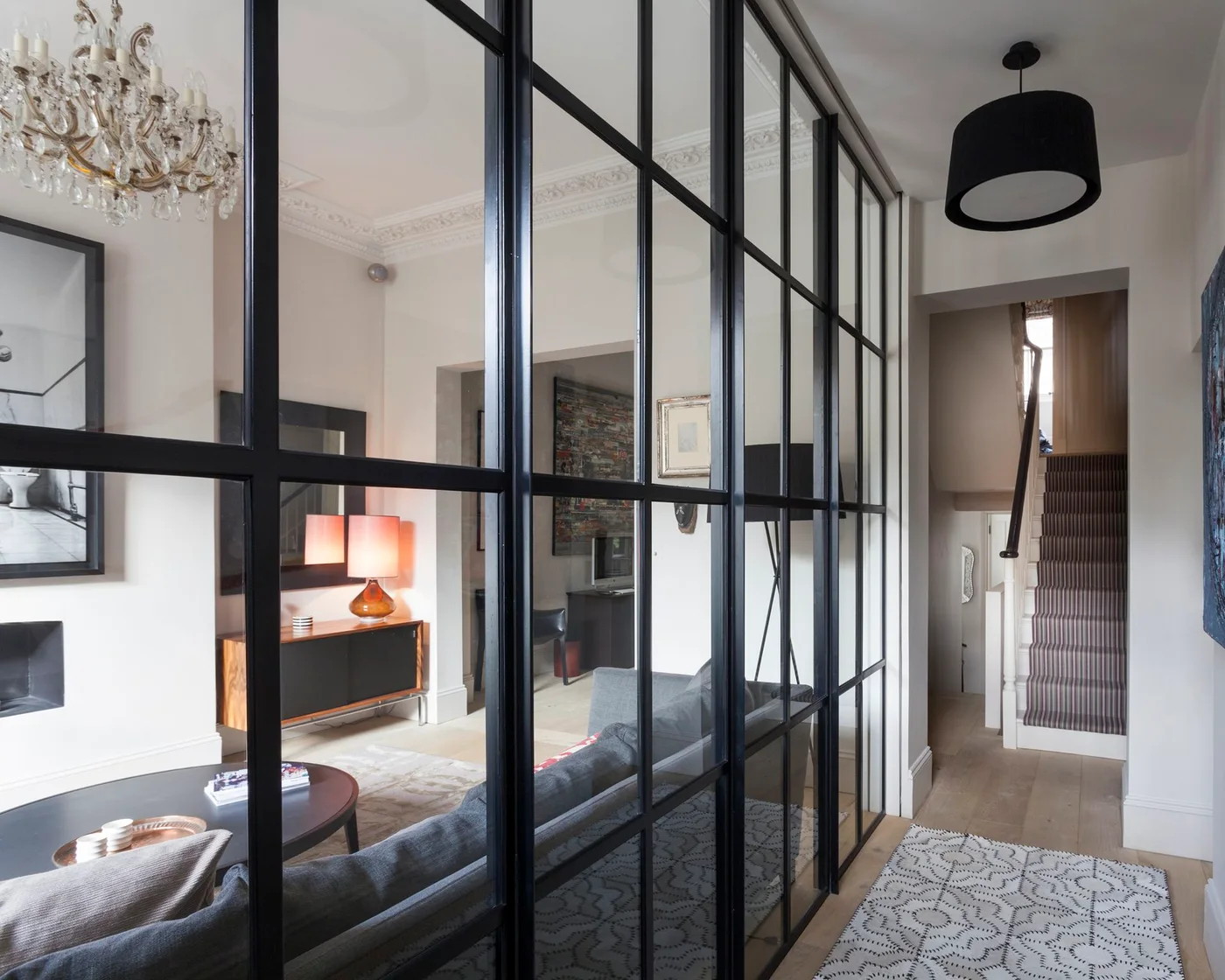 Dark grid-framed glazing divides a pale hallway from a sitting room beside a dark pendant, pale patterned runner and stair doorway