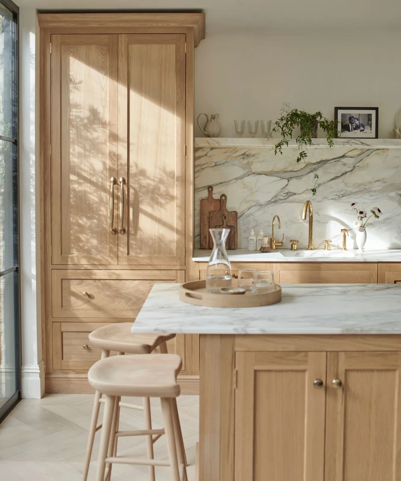 Pale wooden cabinets and stools stand beside a light veined backsplash, pale counter, gold-toned tap and narrow shelf