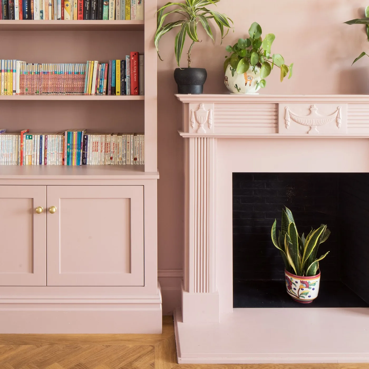 Pink fitted bookcase with lower cupboard beside pink ornate mantel and plant-filled inactive opening