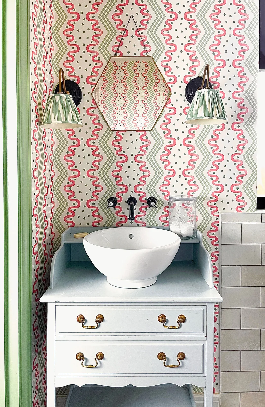 Pink-and-green patterned wallpaper surrounds a small pale vanity and white vessel basin beneath a hexagonal mirror and green patterned shades