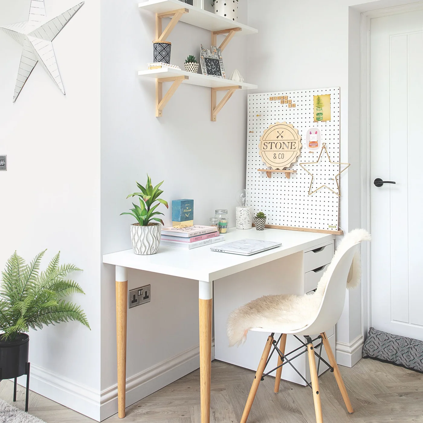 A white desk with wooden legs and a white chair sits in a pale gray corner beneath white shelves and beside a pegboard