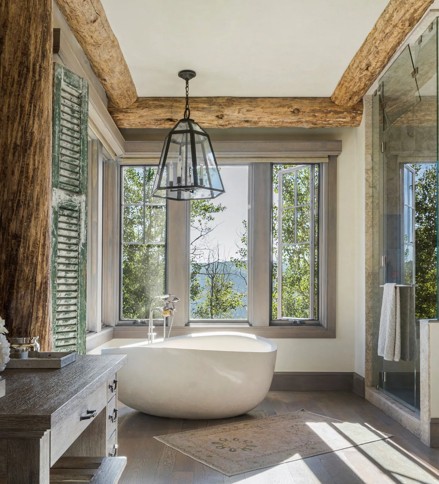 Pale oval bath beneath a large window with exposed wood ceiling beams and a glass shower enclosure