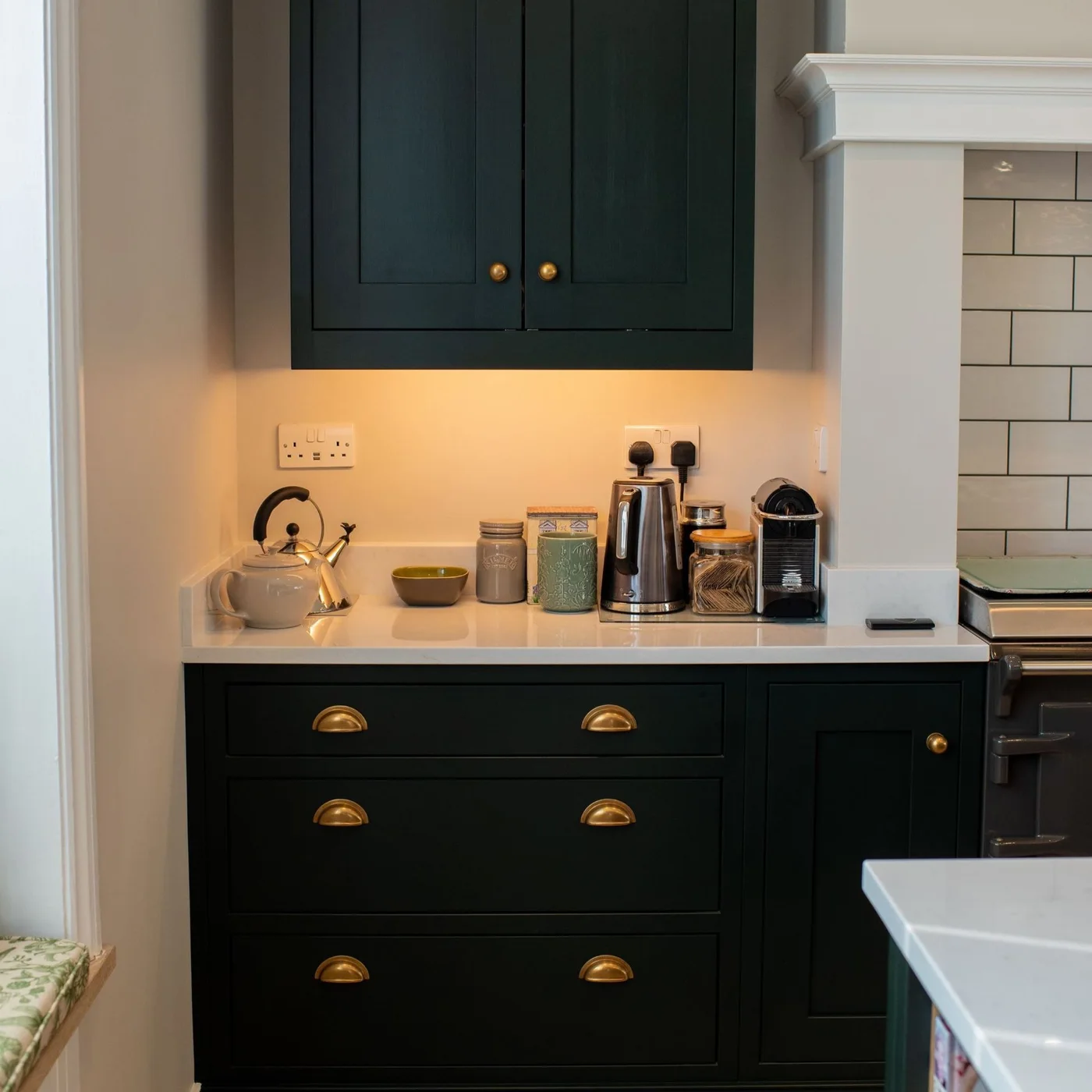 Dark green upper cupboards and lower drawers surround a pale counter with a kettle, coffee machine and jars beneath warm task lighting