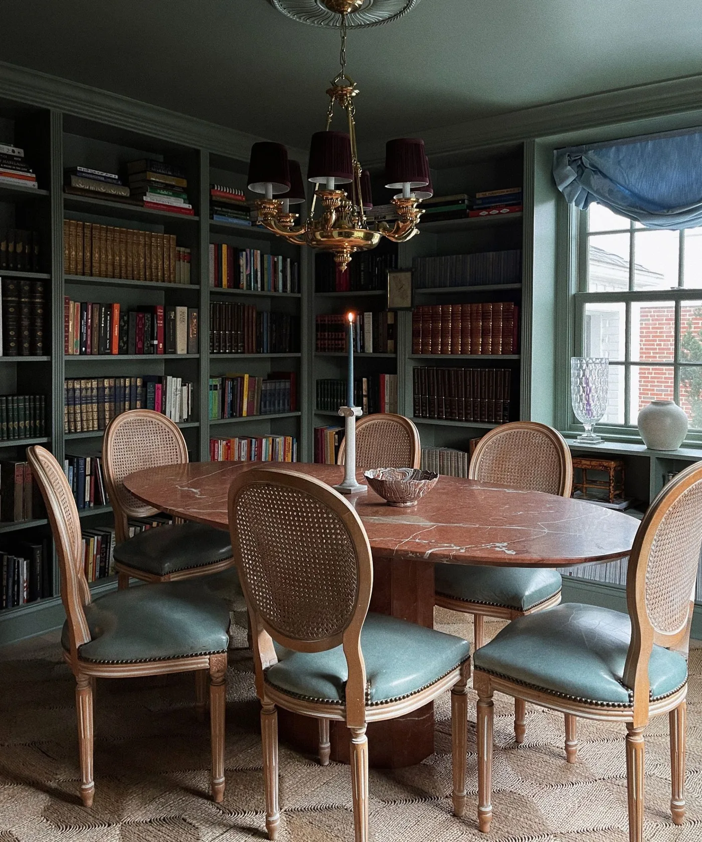Cottage dining room with green bookcases, a dark veined oval tabletop, wooden cane-back chairs with green seats and a shaded ceiling light.