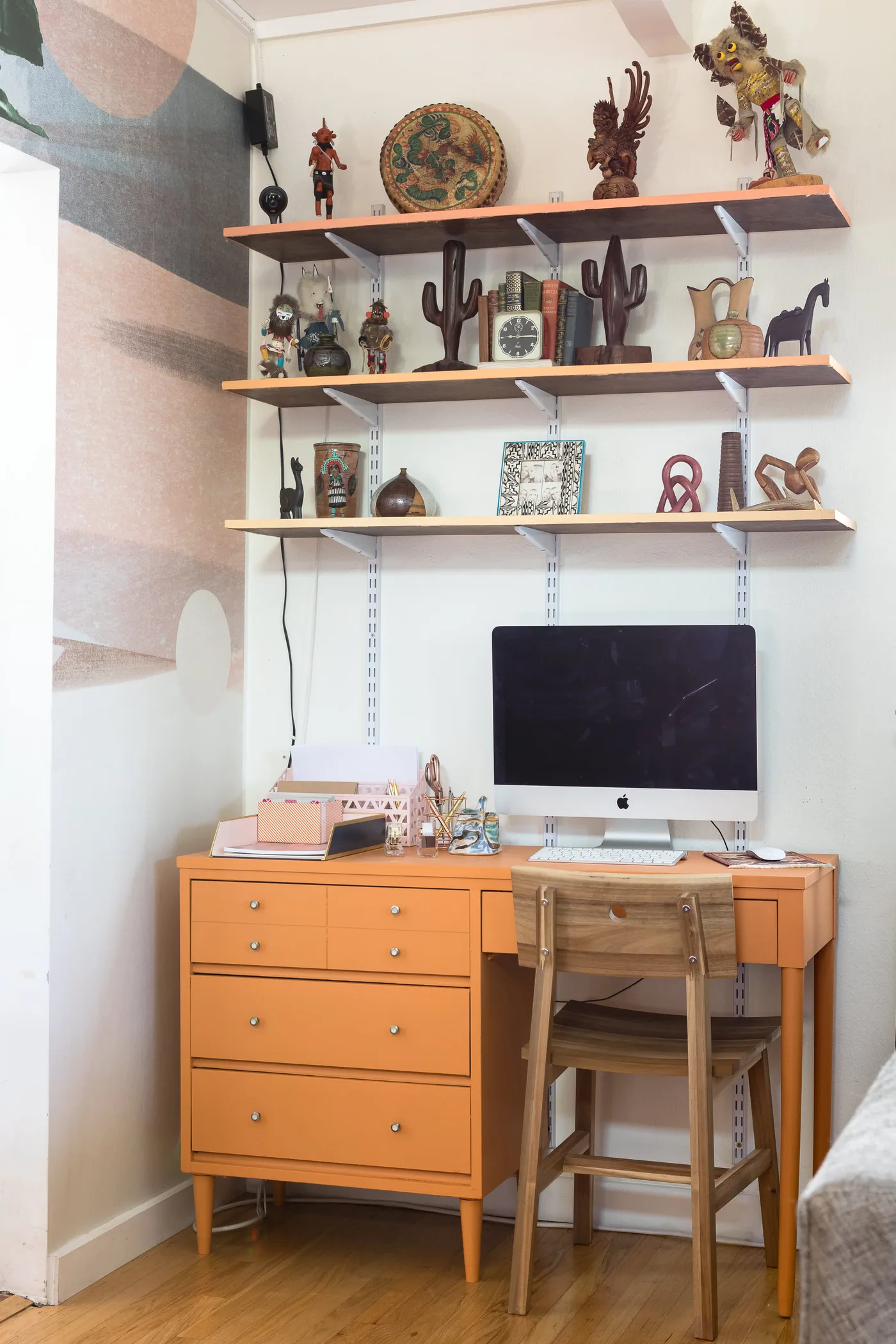 Home workspace with orange drawer desk, wooden chair, computer, three wooden shelves holding books and decorative objects, and multicolored abstract wall at the side.