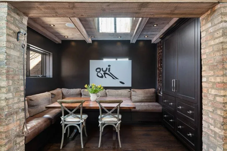Dark full-height cabinets with lower drawers beside a cushioned corner dining booth, wooden table, pale cross-back chairs, brick walls, and a skylight.