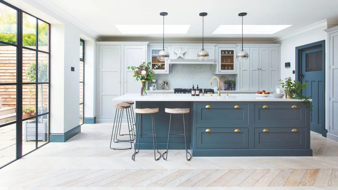 A dark blue island with warm-colored handles and a pale counter stands before lighter blue-gray cupboards below glass pendants and skylights