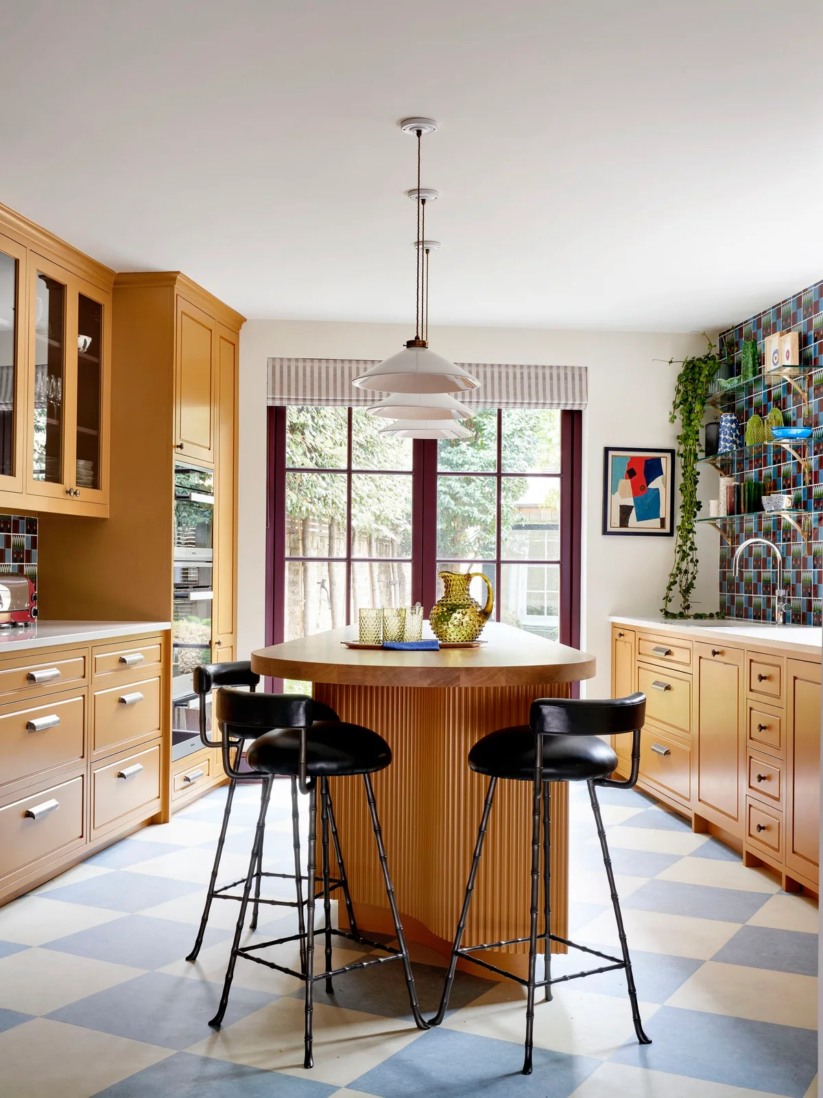 Yellow cabinetry surrounds a round wood-toned island beside multicolored small tiles, purple-red window trim and a pale blue-and-white checked floor