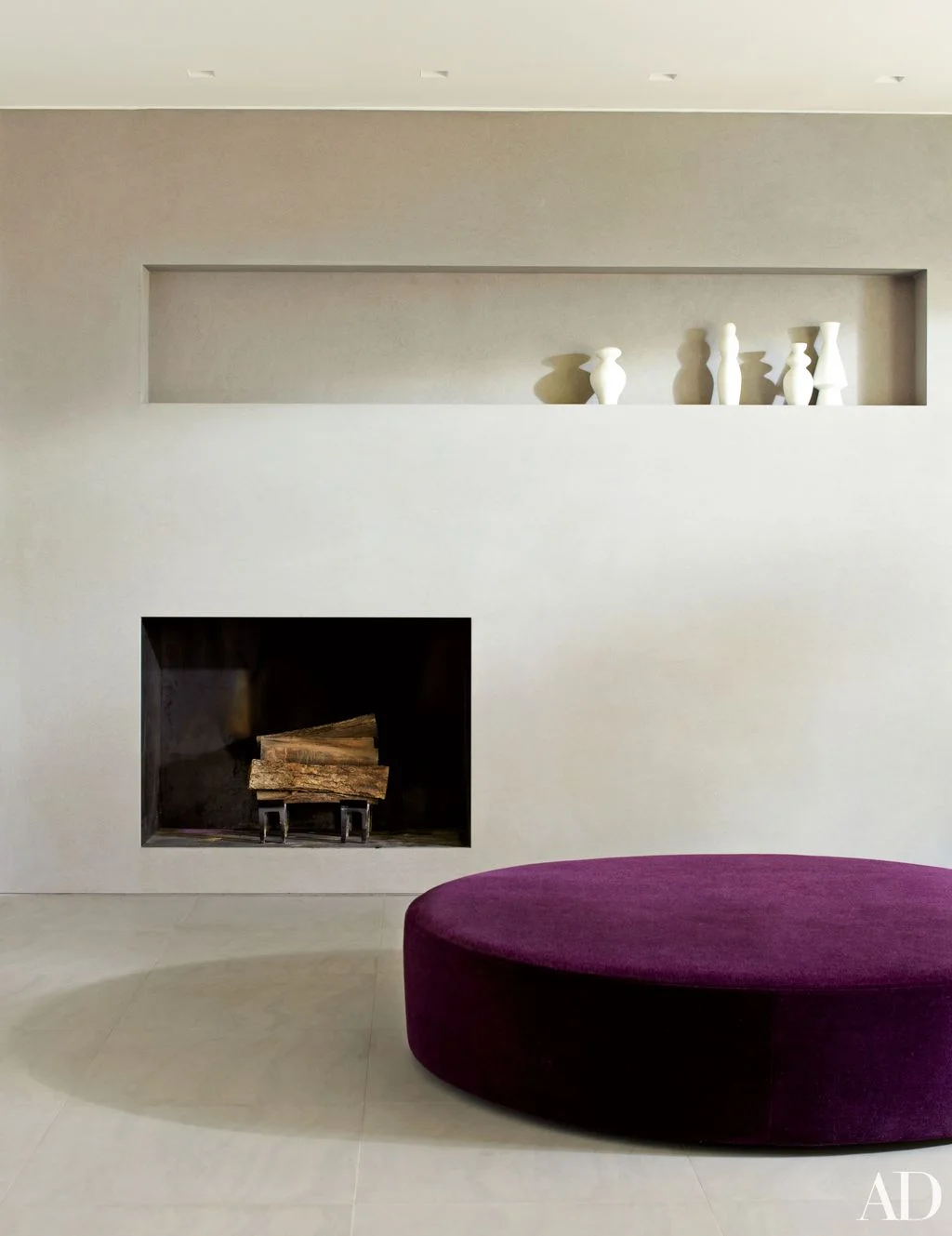 A plain pale fireplace wall contains a dark rectangular opening and a long display recess above a large purple ottoman