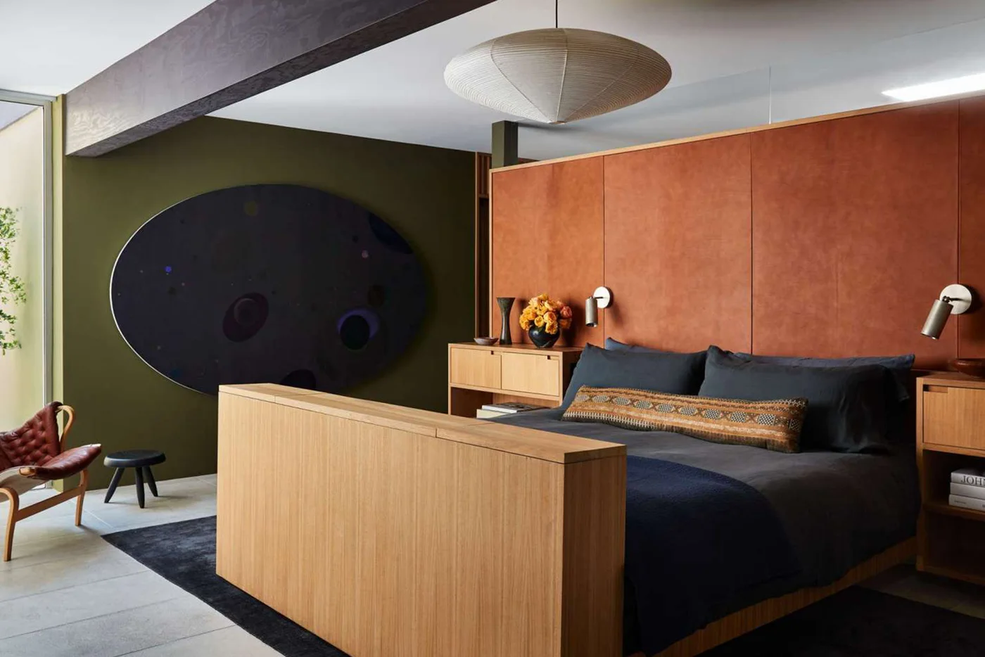 A broad wood bedroom footboard stands before dark bedding beneath a warm orange headboard wall beside pale bedside cabinets, a green wall and rounded pendant