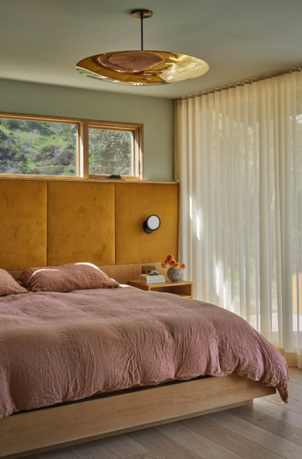 A mustard bedroom headboard sits beneath high wood-framed windows against sage walls beside pink bedding, pale curtains and a broad warm metal-colored pendant