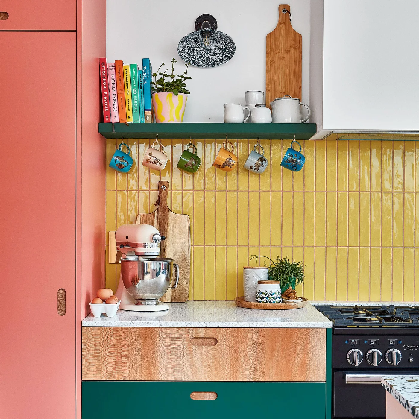 Glossy yellow tiles stand vertically behind a kitchen counter with hanging mugs, green shelving and coral-colored storage