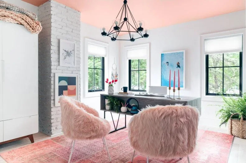 Home office with pink ceiling and patterned rug, gray wooden desk, pale storage cabinet, pink textured guest chairs, black pendant and plants in woven containers.