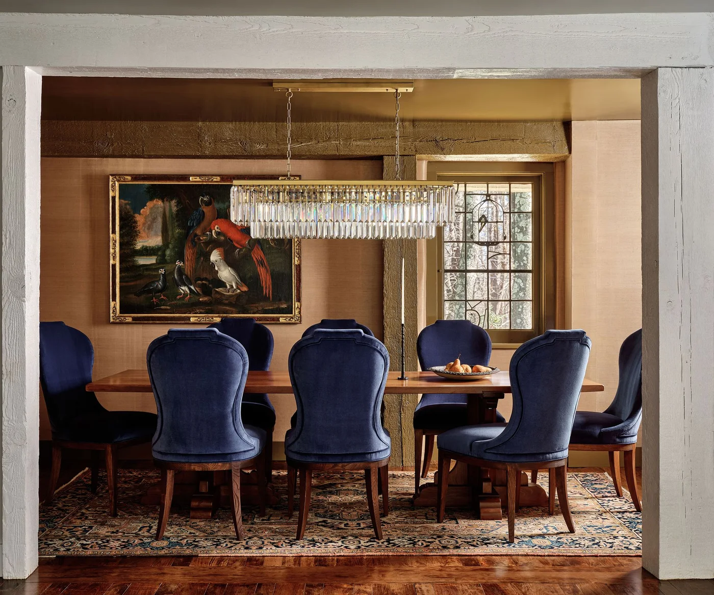 Blue scalloped dining chairs surround a wooden table above a dense patterned rug beneath a long tiered light beside framed bird artwork