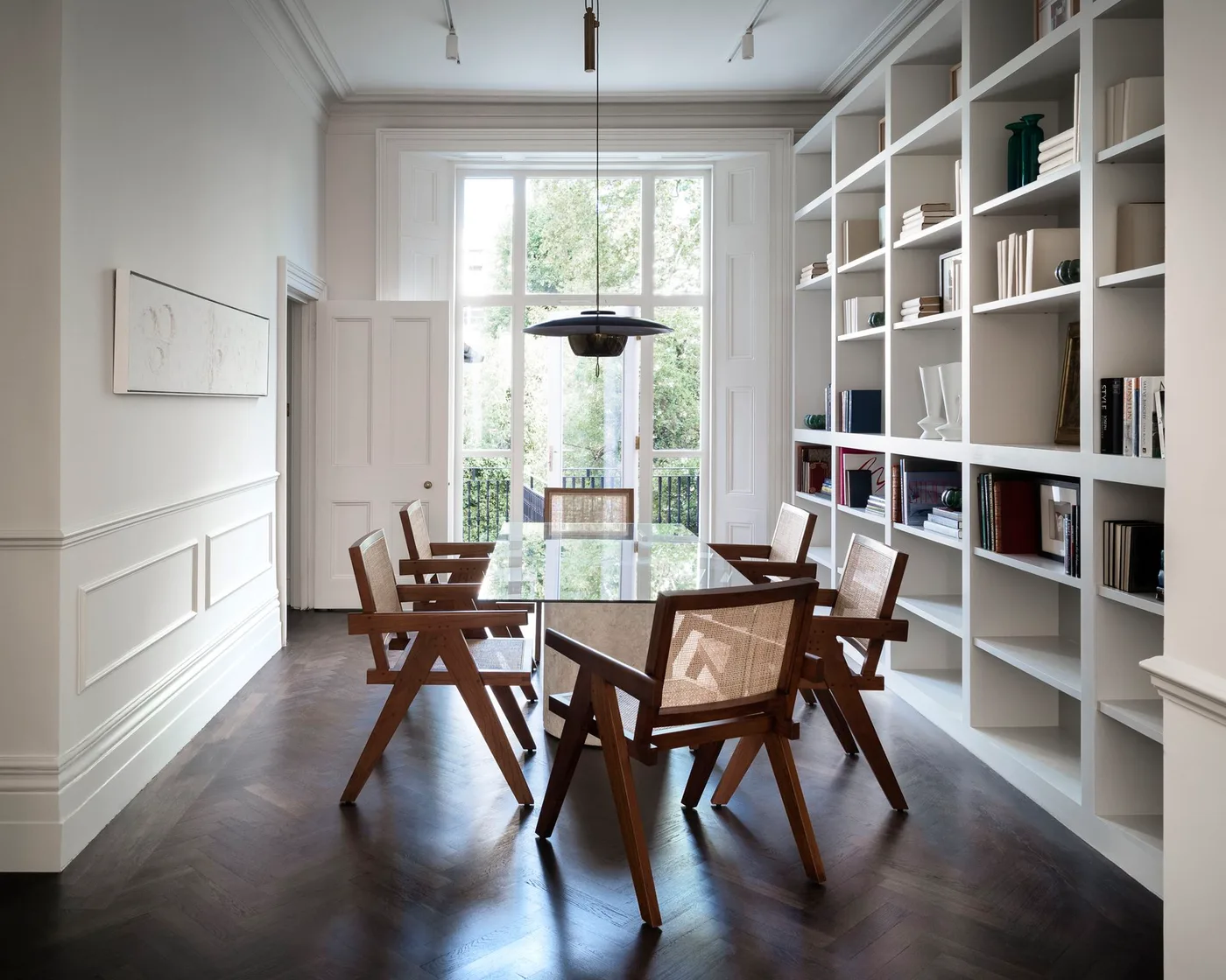 Tall white open shelving beside a long dining table and wood chairs with woven backs near French windows