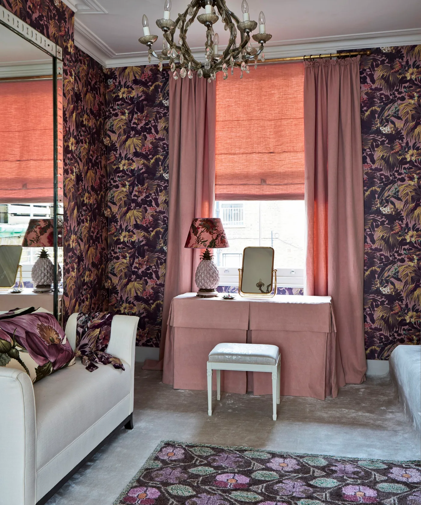 Bedroom with dark floral wallpaper, pink fabric-covered dressing table, pale stool, pink curtains and blind, large mirror and ornate chandelier.