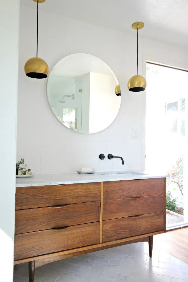 Warm wood vanity with broad drawers beneath a round mirror and two gold-toned pendants