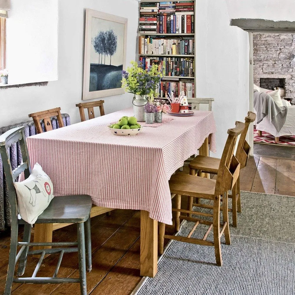 Tall recessed bookcase packed with books beside a rustic dining table with a pink-striped cloth, mixed wooden chairs, pale walls, and a broad doorway.