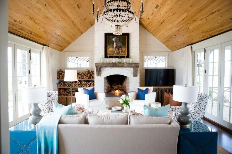 White arched fireplace with thick mantel and picture beneath pitched wood ceiling, with log shelves and bright seating