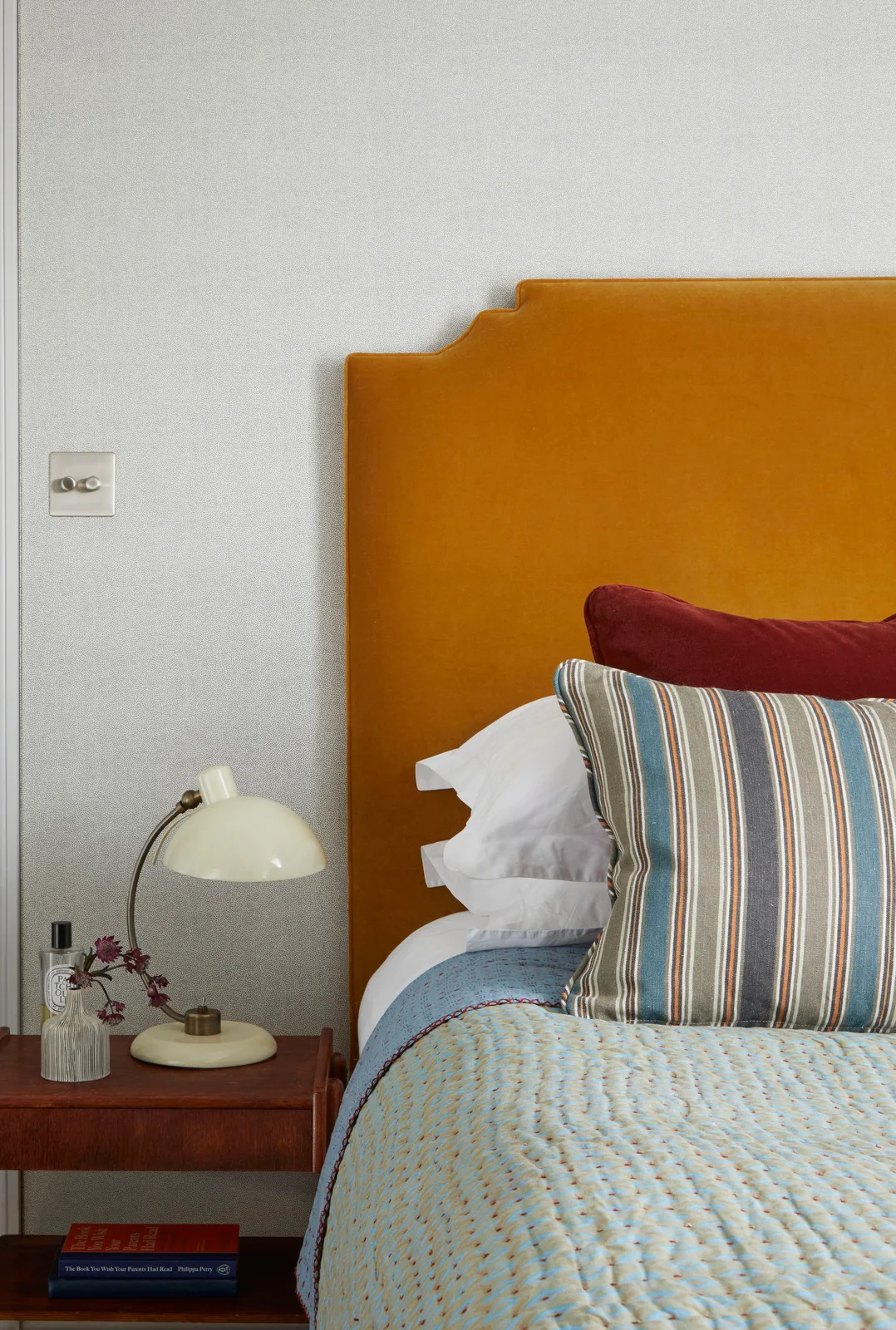 An orange bedroom headboard sits beside a wood bedside table and pale rounded lamp behind striped cushions, a burgundy pillow and light blue bedding
