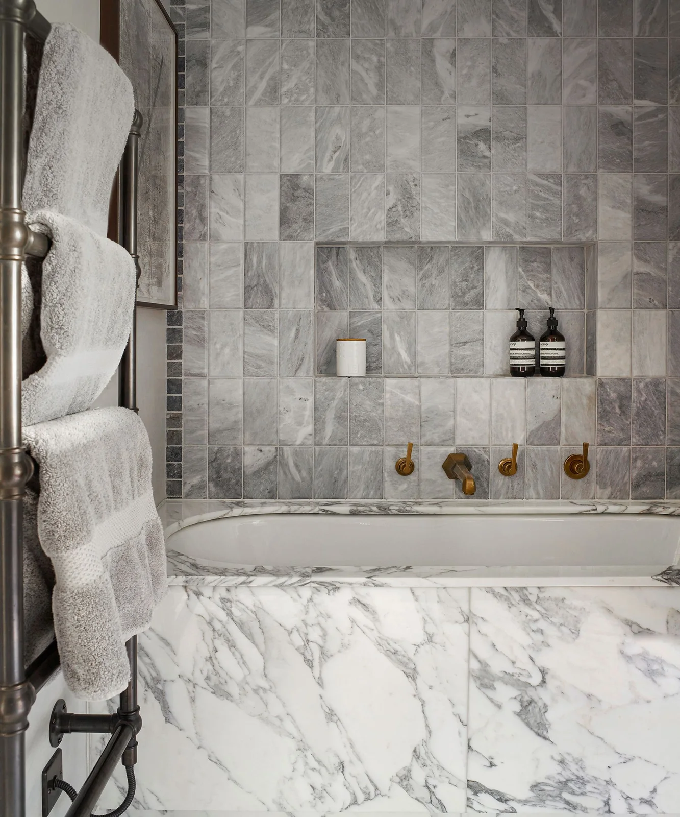 A white inset bath with a pale veined surround sits beneath gray veined square wall tiles and a recessed shelf holding bottles beside a towel rail