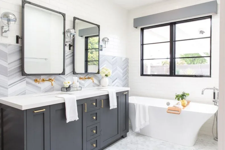 Bathroom with charcoal double vanity, warm metal handles, white countertop, pale chevron backsplash, dark framed mirrors, globe wall lights, and white tub.
