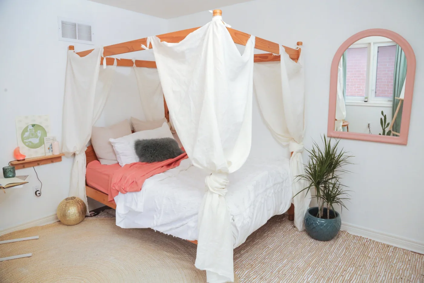 Student bedroom with a wooden four-post bed, loose white canopy fabric, coral and white bedding, pink framed mirror and a tall plant in a blue pot.
