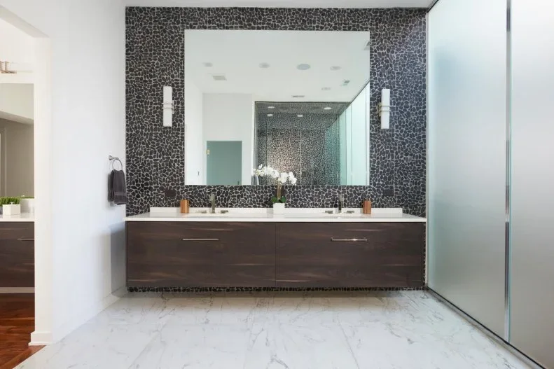A dark floating vanity with two basins sits below a broad mirror surrounded by a small dark stone-like wall pattern beside pale flooring and frosted glass