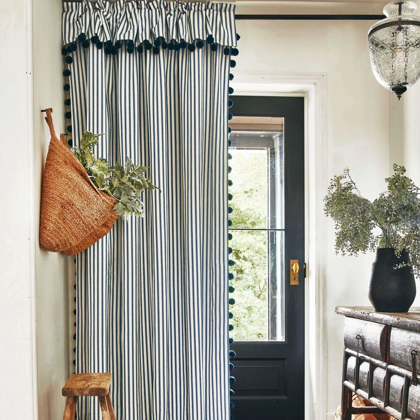 Country entrance with a dark glazed door, long blue striped curtain with decorative edging, wooden console and woven bag hanging on the pale wall.