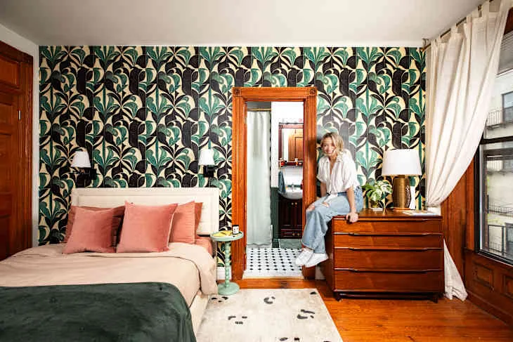 Bedroom with green botanical wallpaper, pale bed and pink pillows, green throw, warm wooden dresser and large wood-framed mirror.