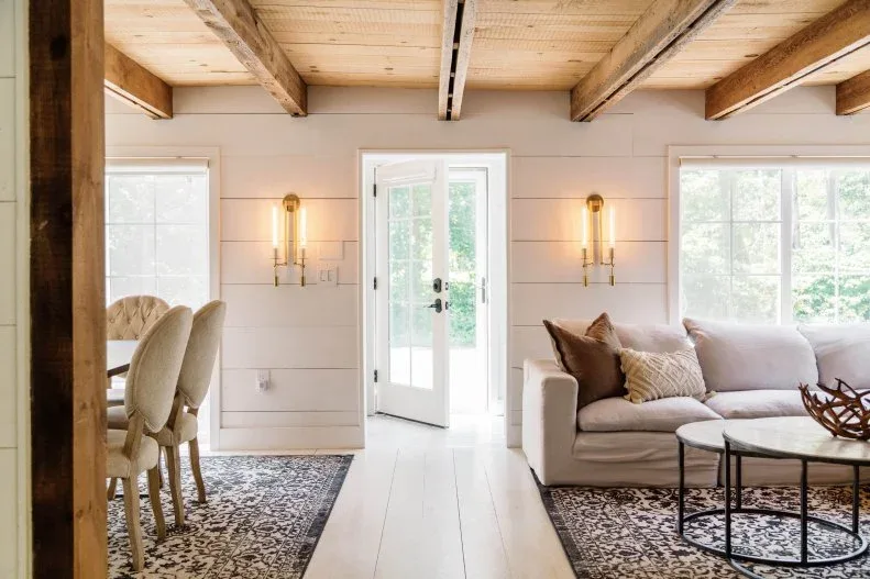 Open farmhouse entrance with glazed door, white horizontal paneling, wood beam ceiling, paired wall lights and pale living-room seating