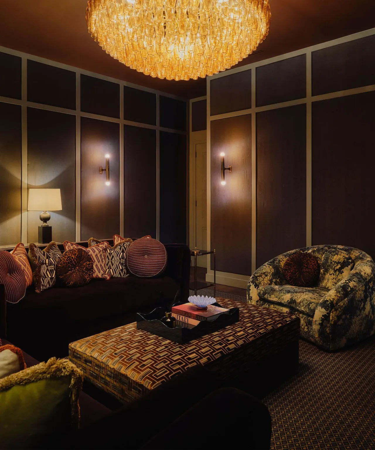 Dark paneled room with a sofa, patterned chair and upholstered patterned ottoman beneath warm lighting