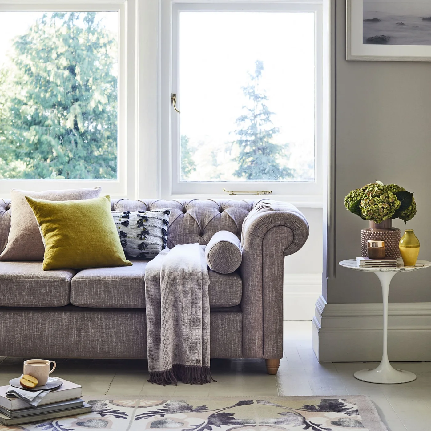 A gray tufted sofa with a yellow cushion sits beside a small pale pedestal table and a floral rug