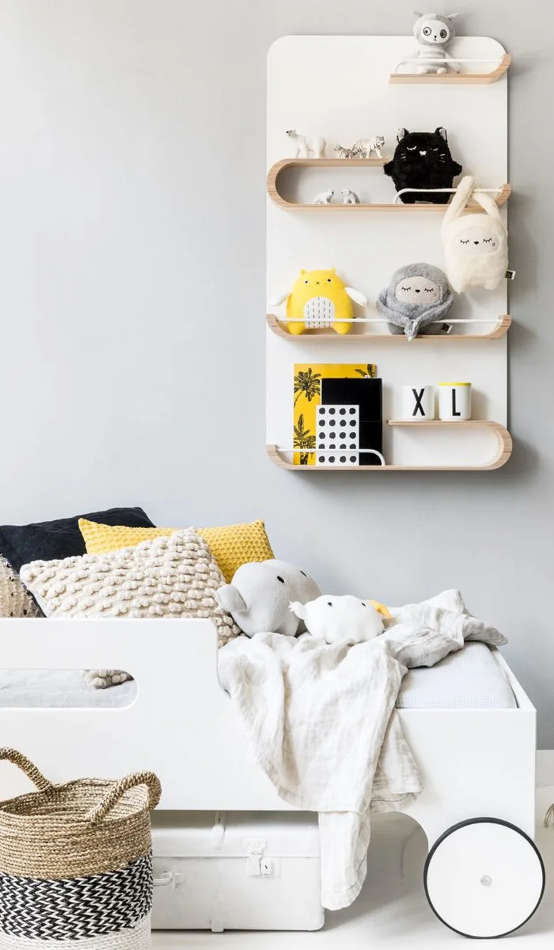 A rounded pale wood wall shelf holds a few toys and books above a white child’s bed with yellow and black cushions and woven baskets nearby