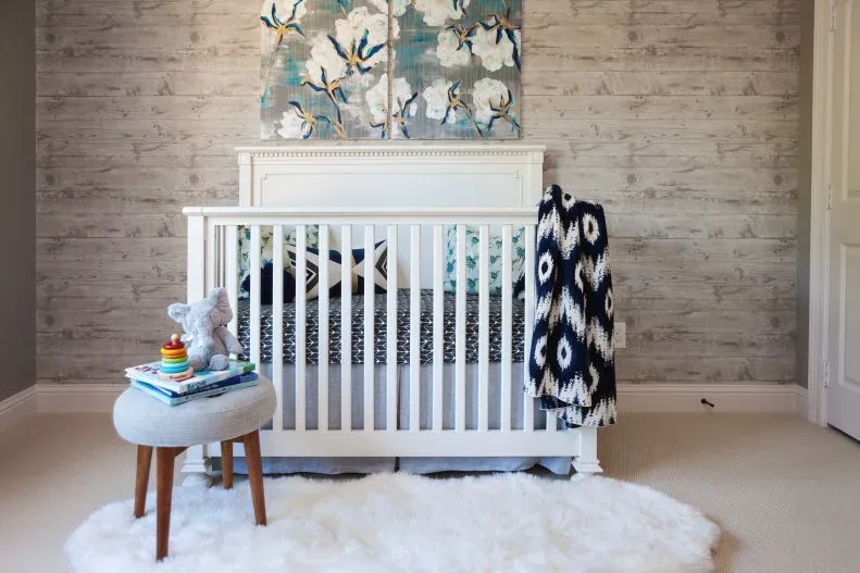 Pale paneled crib beneath floral picture with quiet textured-looking wall, pale stool and dark staged throw.