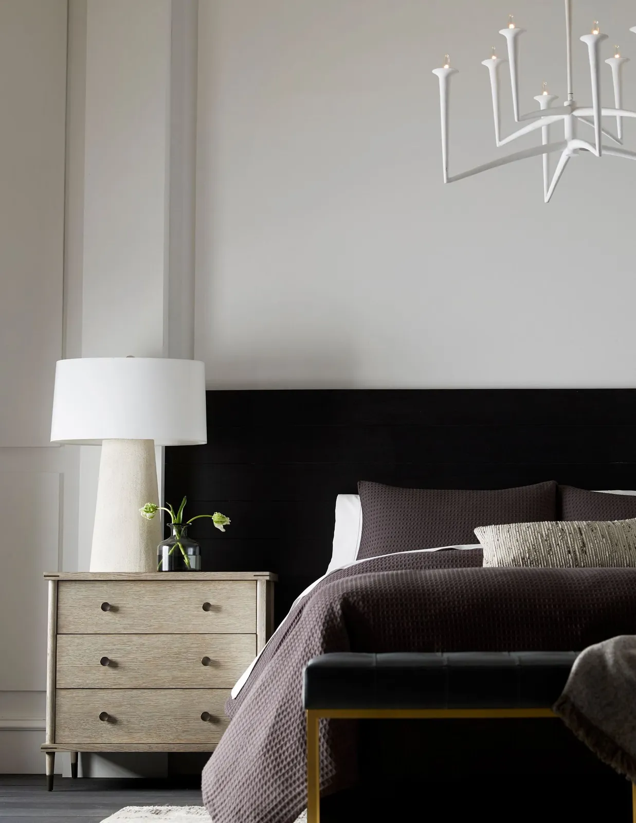 A dark headboard sits against pale walls beside a compact drawer unit and lamp.