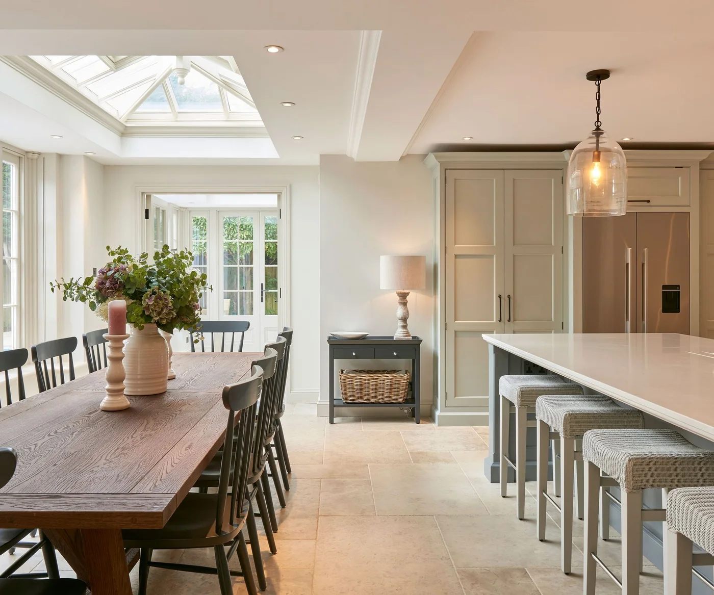 A long wooden table and dark chairs stand parallel to a pale kitchen island beneath a glazed roof light