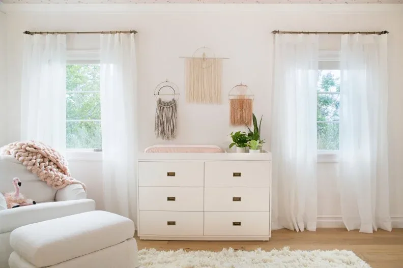A white dresser, pale gray armchair and round pouf sit between windows beneath muted wall hangings