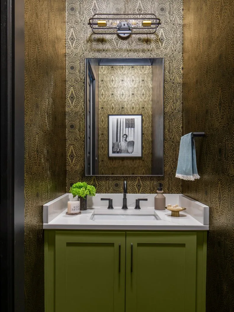 Powder room with fine dark-and-gold geometric wallpaper, olive-green vanity, rectangular dark mirror, black tap, caged wall light and blue hand towel.