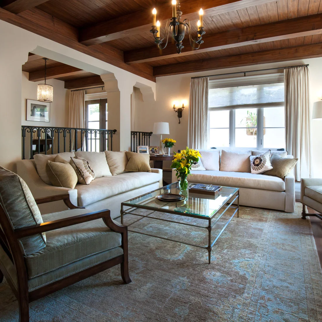 Pale living-room sofas sit beneath dark wooden ceiling beams beside decorative metal railings, a wood-armed chair and a glass-topped coffee table