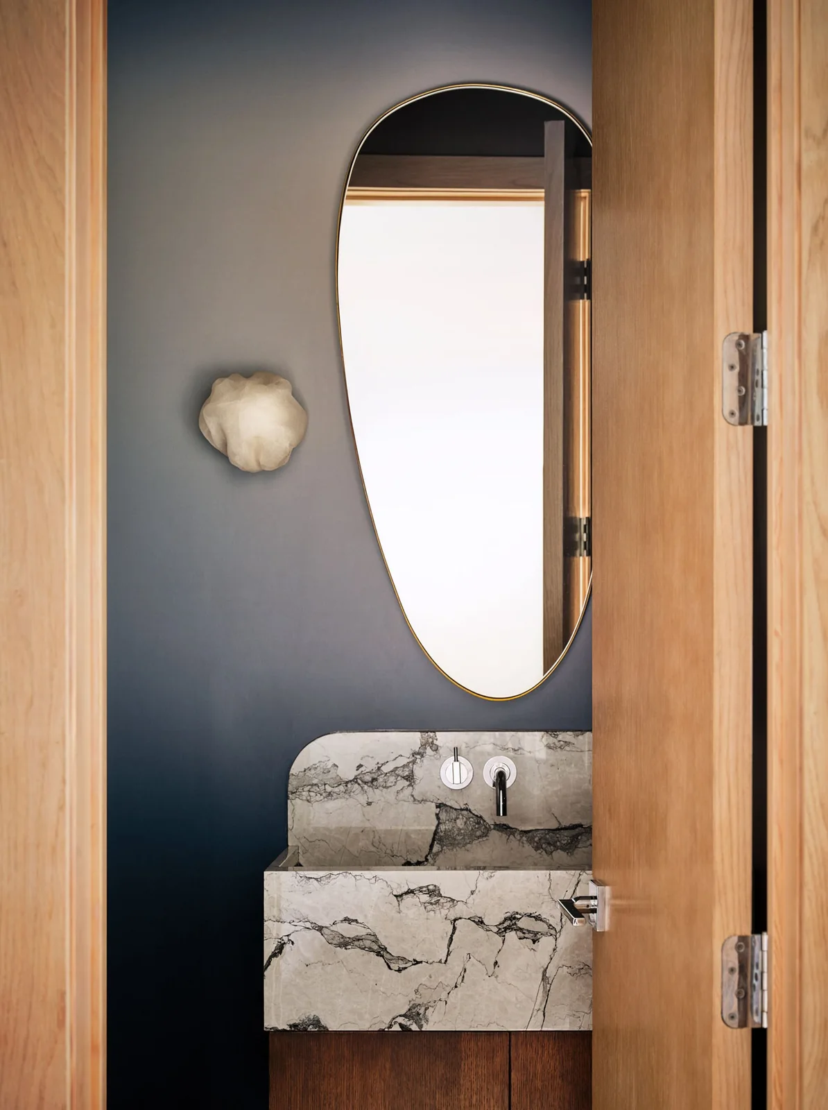 A tall oval mirror hangs over a pale dark-veined basin counter against a blue-gray wall beside a small pale wall light and wood door frames