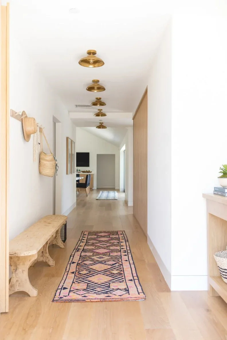 A wooden bench stands below pale wall hooks holding a hat and woven bag beside a patterned runner and tall wooden storage wall