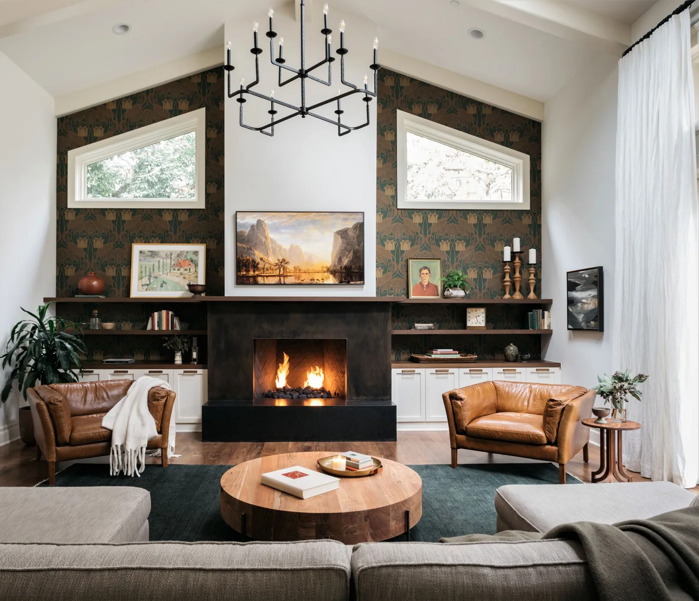 Tall living room fireplace wall with green-and-brown wallpaper, brown armchairs, pale sectional and round wood table