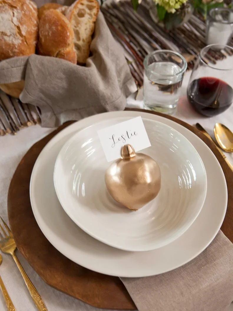 A pale name card sits behind a gold-toned decorative pear in a white bowl above a pale napkin and round charger