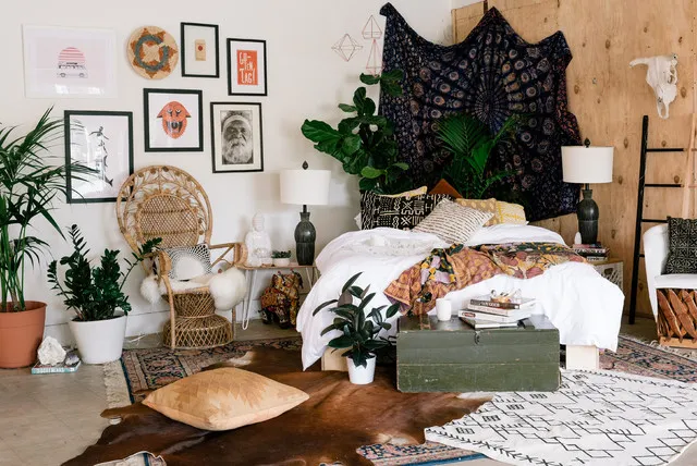 A dark tapestry hangs behind a pale bed among woven furniture and leafy plants.