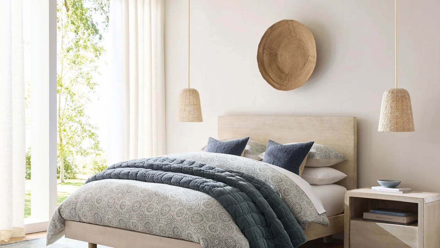 A pale wooden bed with blue-and-white bedding sits beneath two woven pendant shades and a round wooden wall piece beside a light bedside table