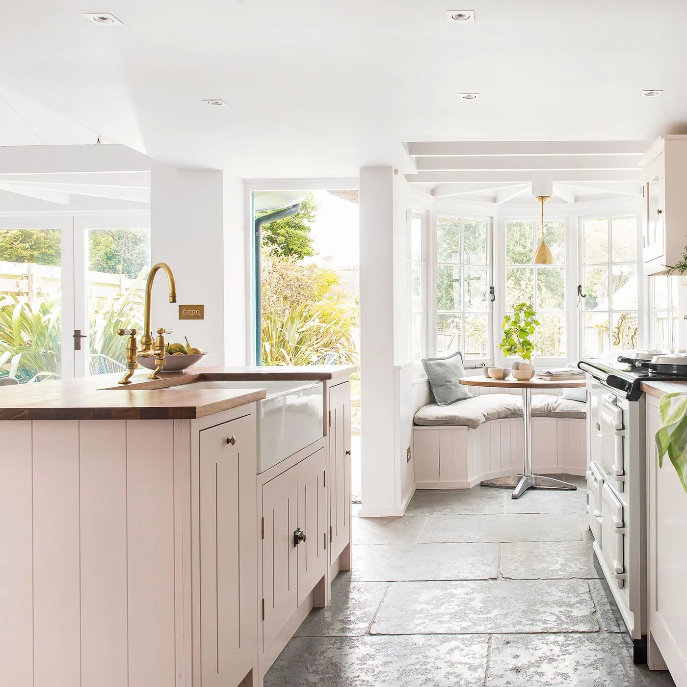 Bright kitchen with pale pink paneled island, white apron-front sink, dark worktop and a cushioned window dining nook.