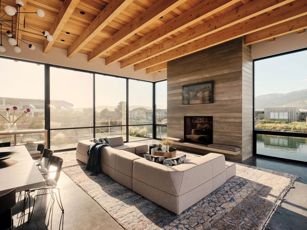 A pale modular sofa sits below a broad timber ceiling beside large windows.